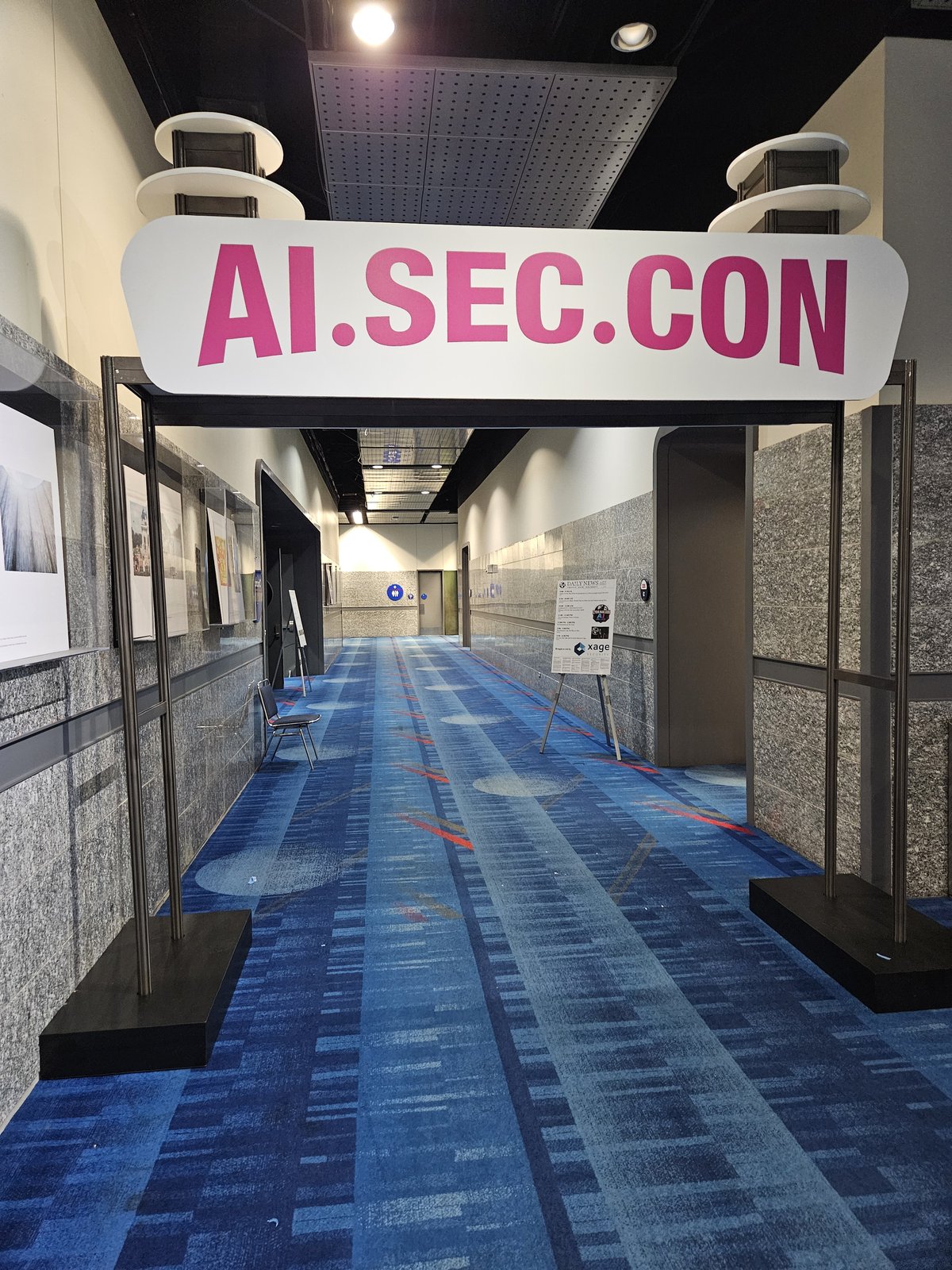 A hot pink illuminated archway reading AI.SEC.CON spanning a convention center corridor, with attendees walking through underneath.