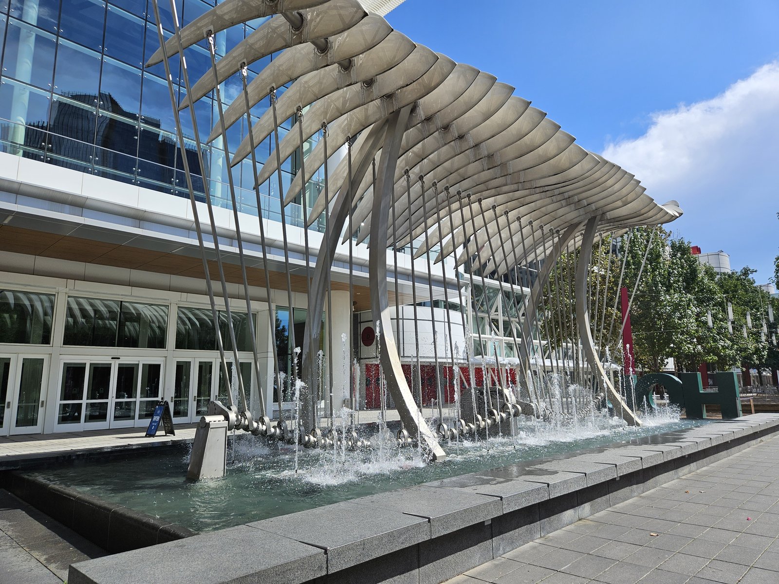 The exterior of the George R. Brown Convention Center on a clear sunny day, a white sail-shaped fin canopy rising over a fountain plaza.