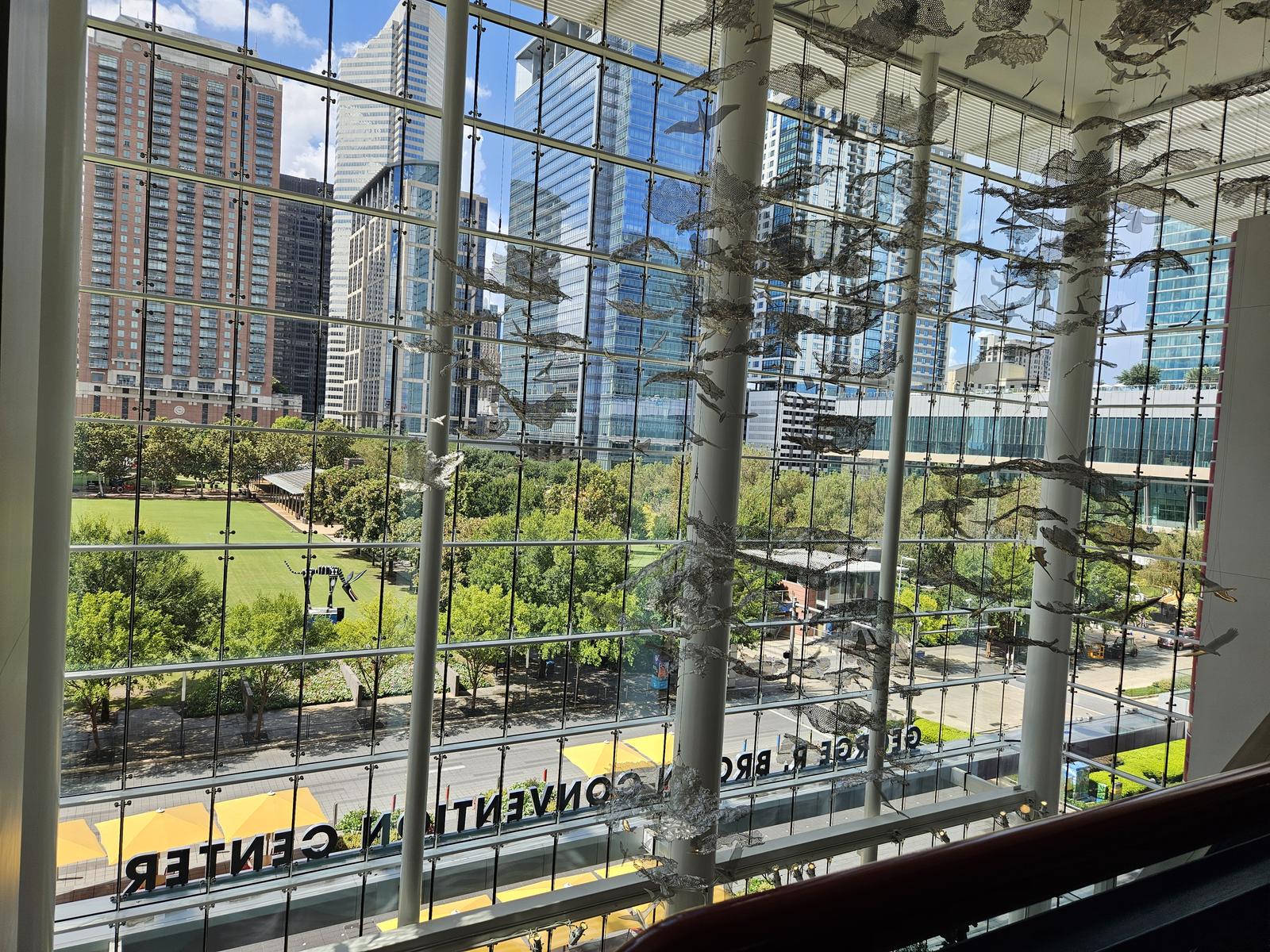 An interior window wall with hanging wire-mesh bird sculptures suspended in front of the glass, Discovery Green park and the Houston skyline visible beyond.