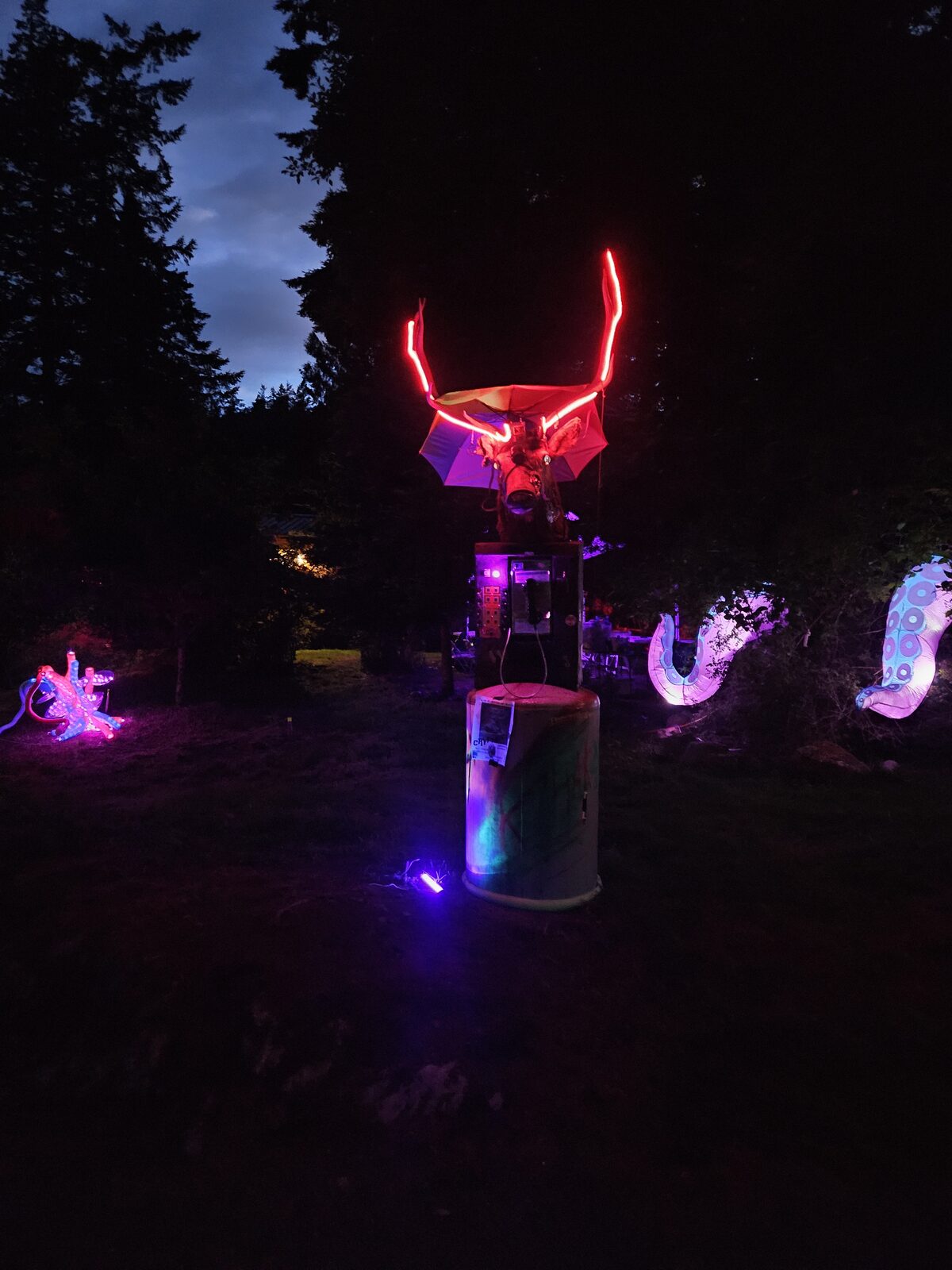 A night art installation, a red-outlined winged figure with hanging LED jellyfish in the trees.