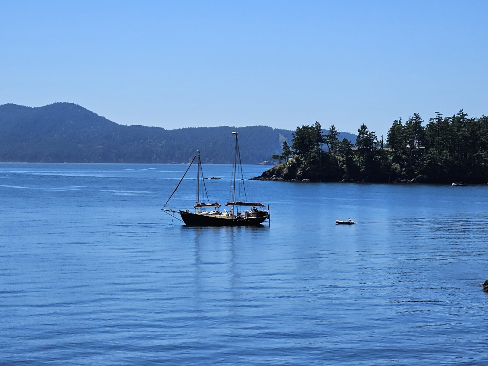 A sailboat anchored in the Sound, a forested island behind, a clear blue day.