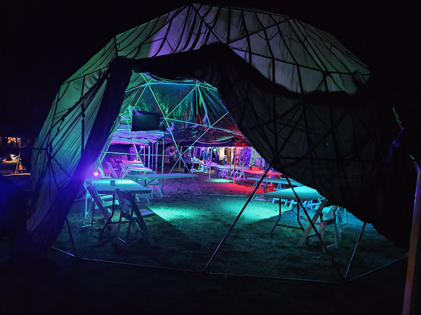 A geodesic dome interior at night, RGB-lit, with tables and chairs and night-market wares in the back.