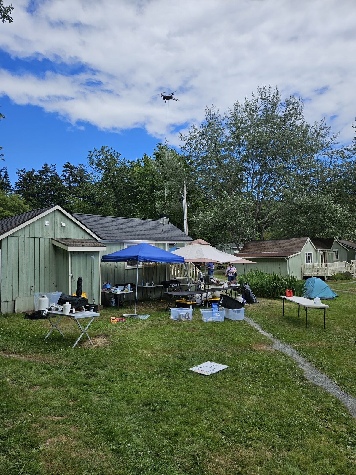A daytime staff and workshop setup on a green house deck, a drone flying overhead in a blue sky.