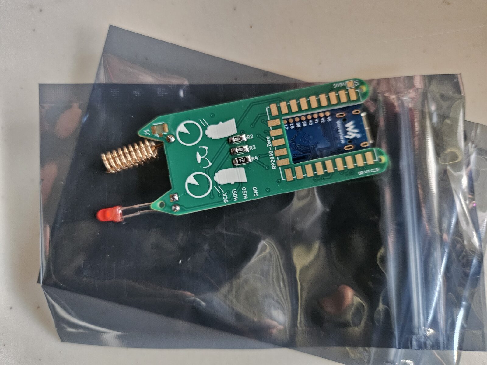 A small circuit board, an RP2040-Zero microcontroller with a helical antenna, sitting in an antistatic bag.