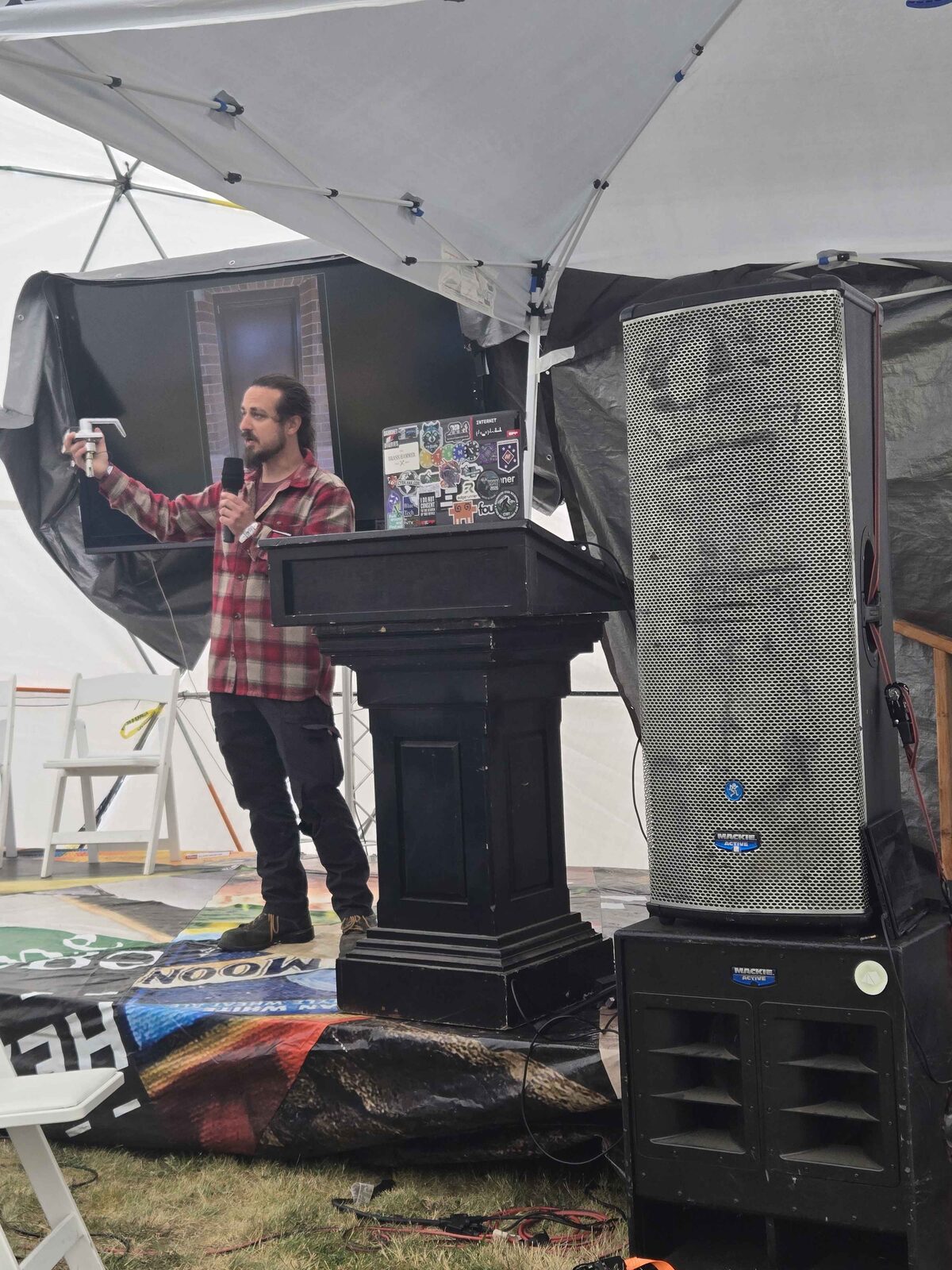 The author at the ToorCamp main-stage podium in a red plaid shirt, holding up an L-shaped lock tool, laptop covered in stickers including a Darknet Diaries sticker.