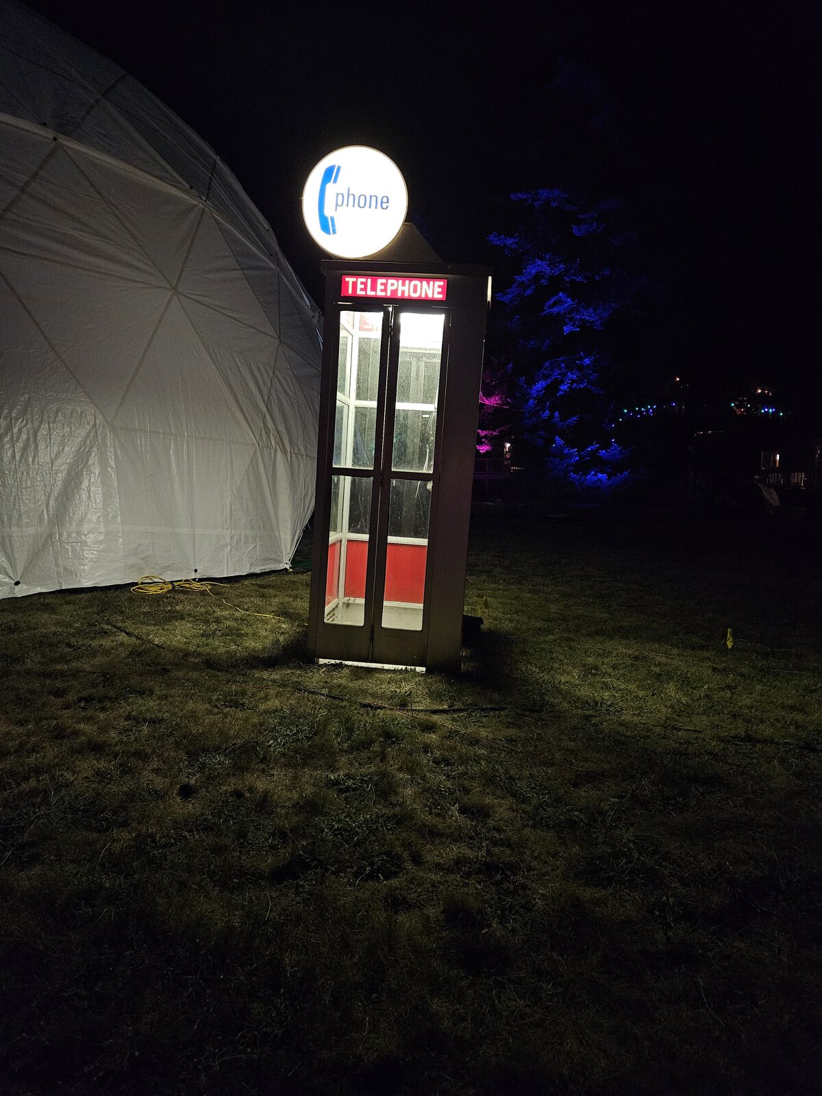 A glowing telephone booth beside a dome tent at night.