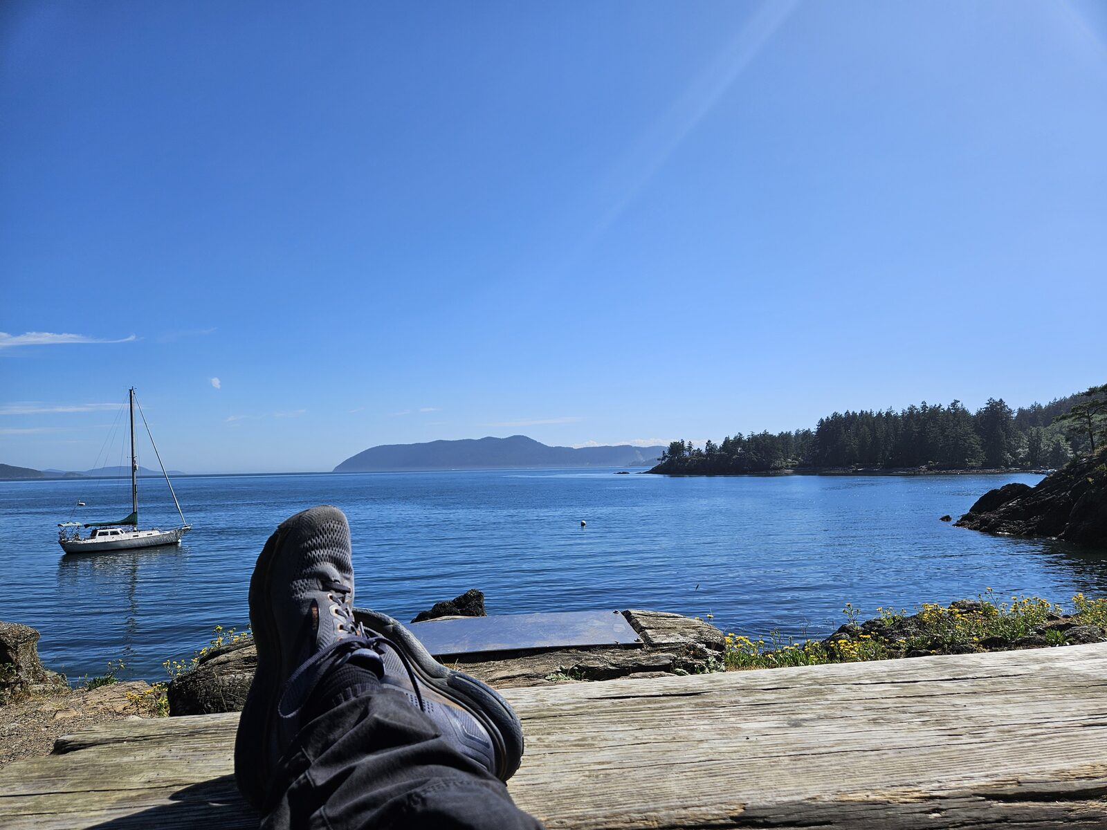 The author's feet up on a log, a sailboat and forested islands and blue water beyond.