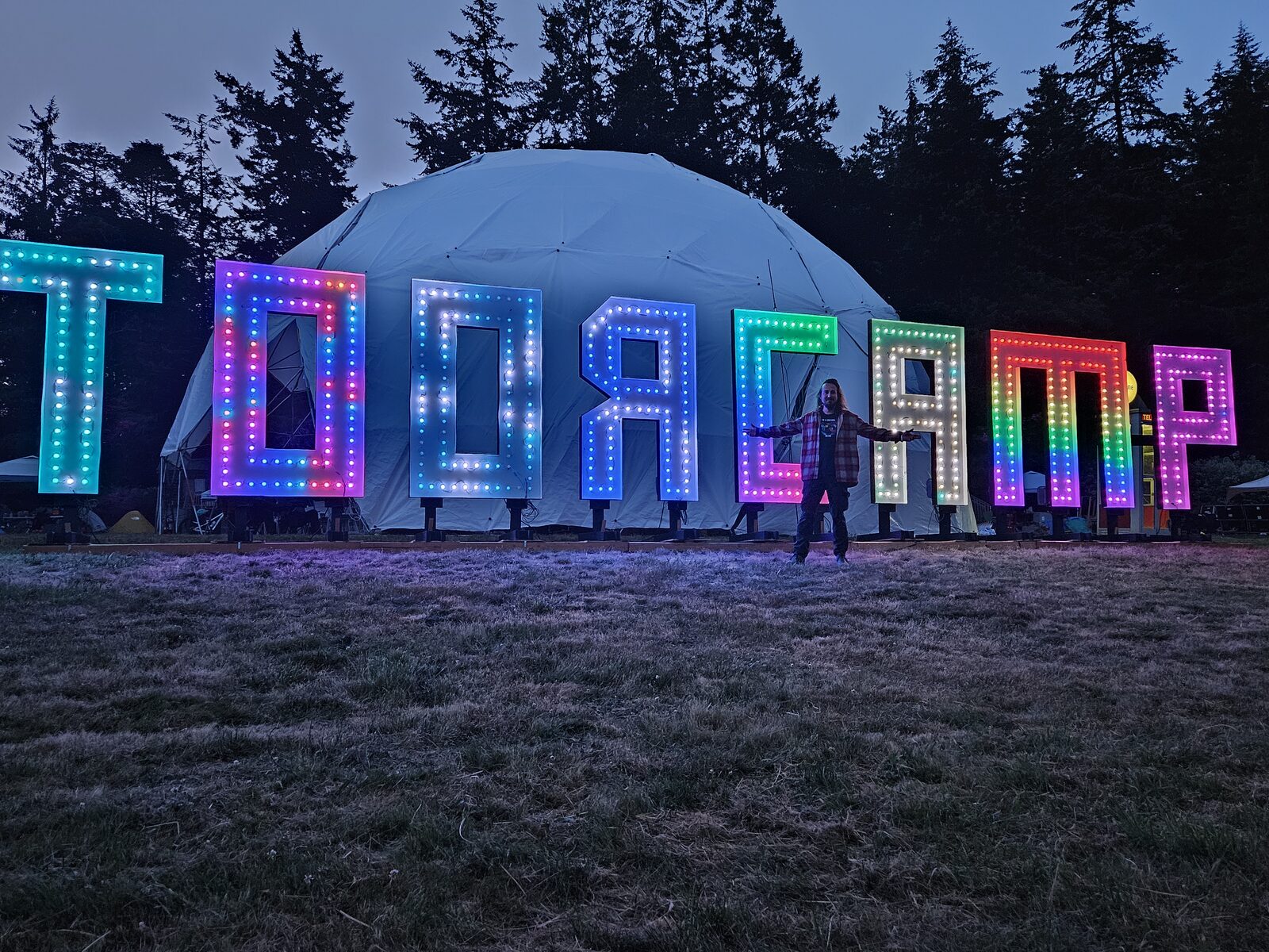 The author standing with arms wide in front of giant illuminated rainbow TOORCAMP marquee letters at dusk, a geodesic dome behind.